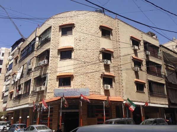 A multi-story building with a stone facade, multiple balconies, and several air conditioning units. Flags are displayed outside the entrance. - Alaamira Furnished Apartments (Chiyah)