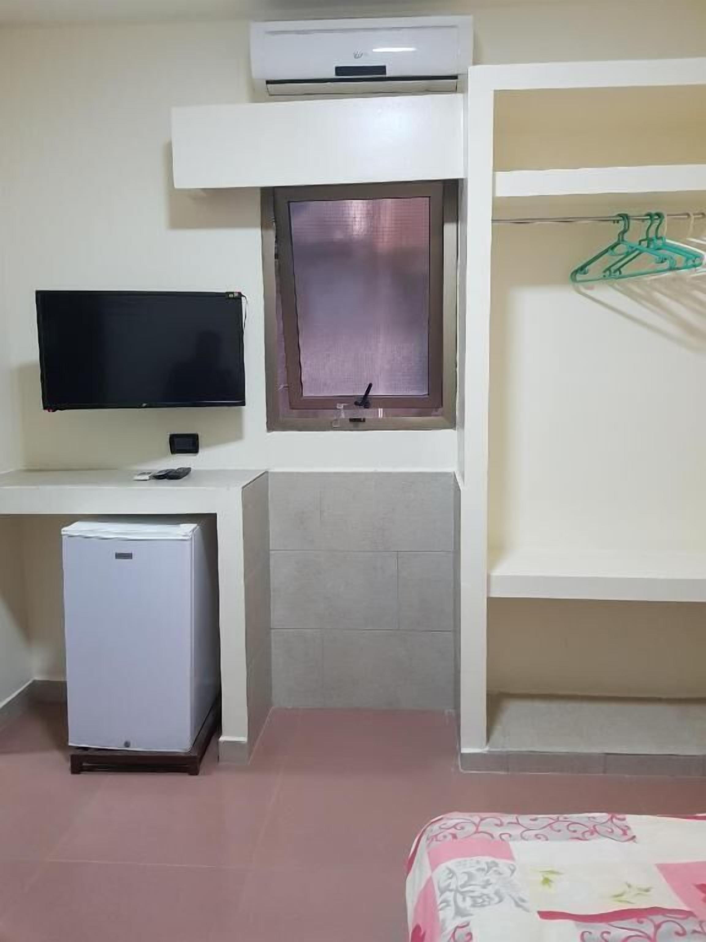 Ein kleines Zimmer mit einem Bett, einem Schreibtisch mit Computer, einem Fernseher und einem Fenster mit Vorhang.