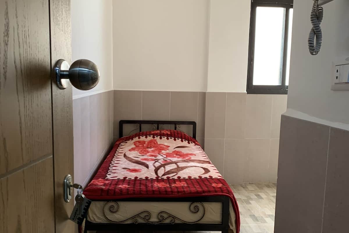 A small, narrow room with a bed covered in a floral-patterned blanket, a wooden door, and a window with a telephone on the wall.