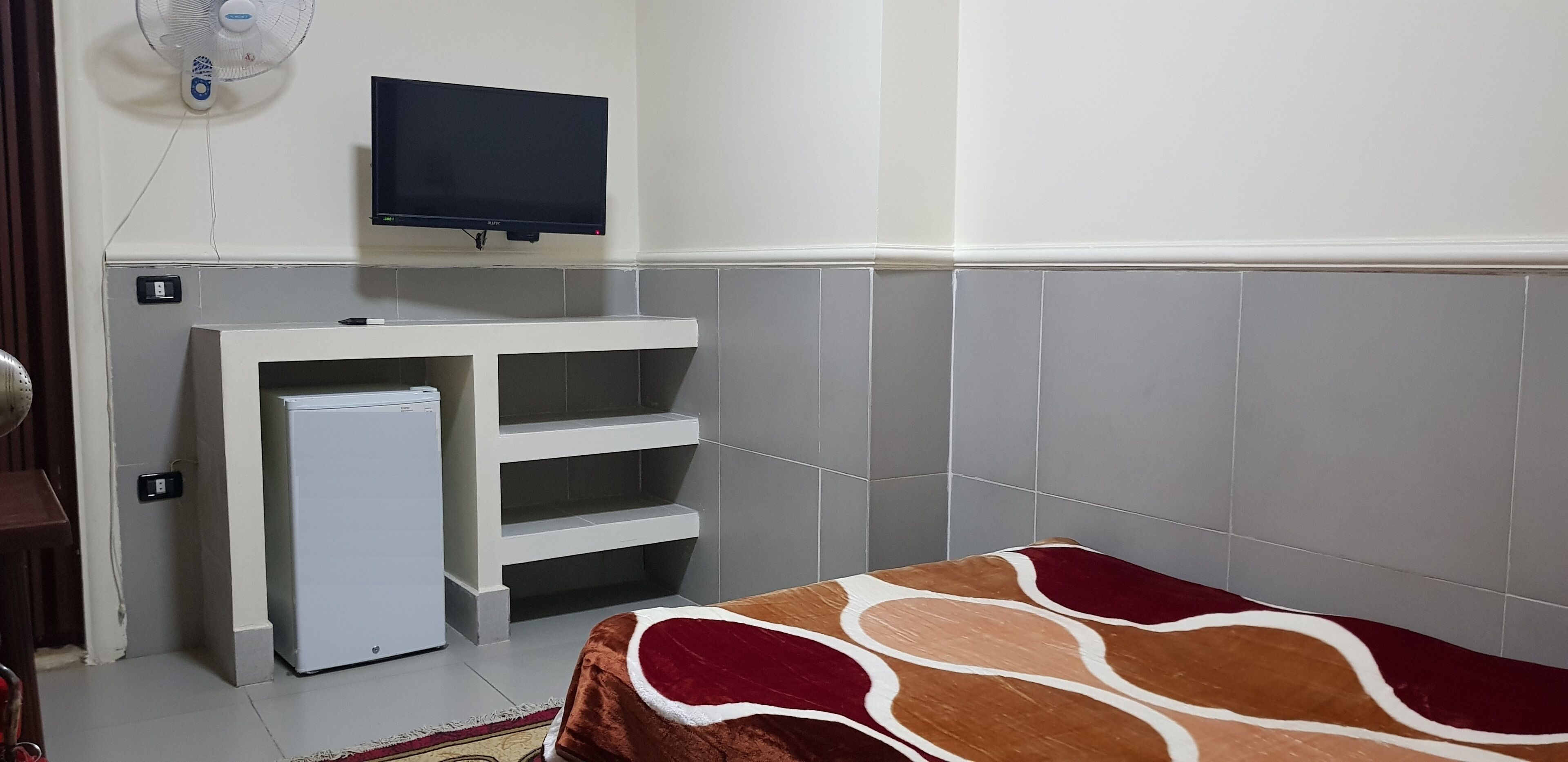 A small, clean room with a bed, a TV mounted on the wall, a small fridge, and a heater.