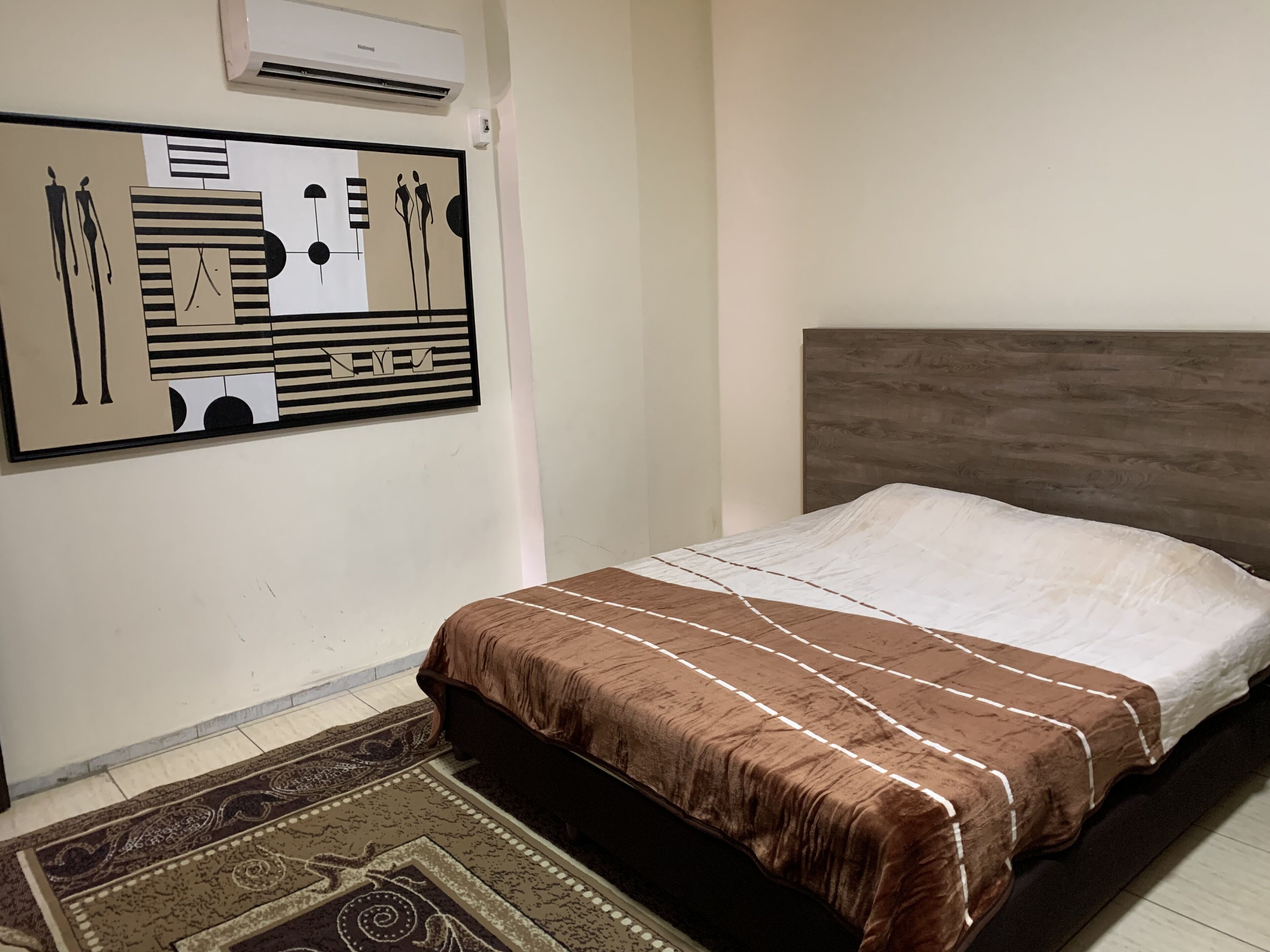 A bedroom with a bed, a headboard, a wall-mounted air conditioner, and a patterned rug.