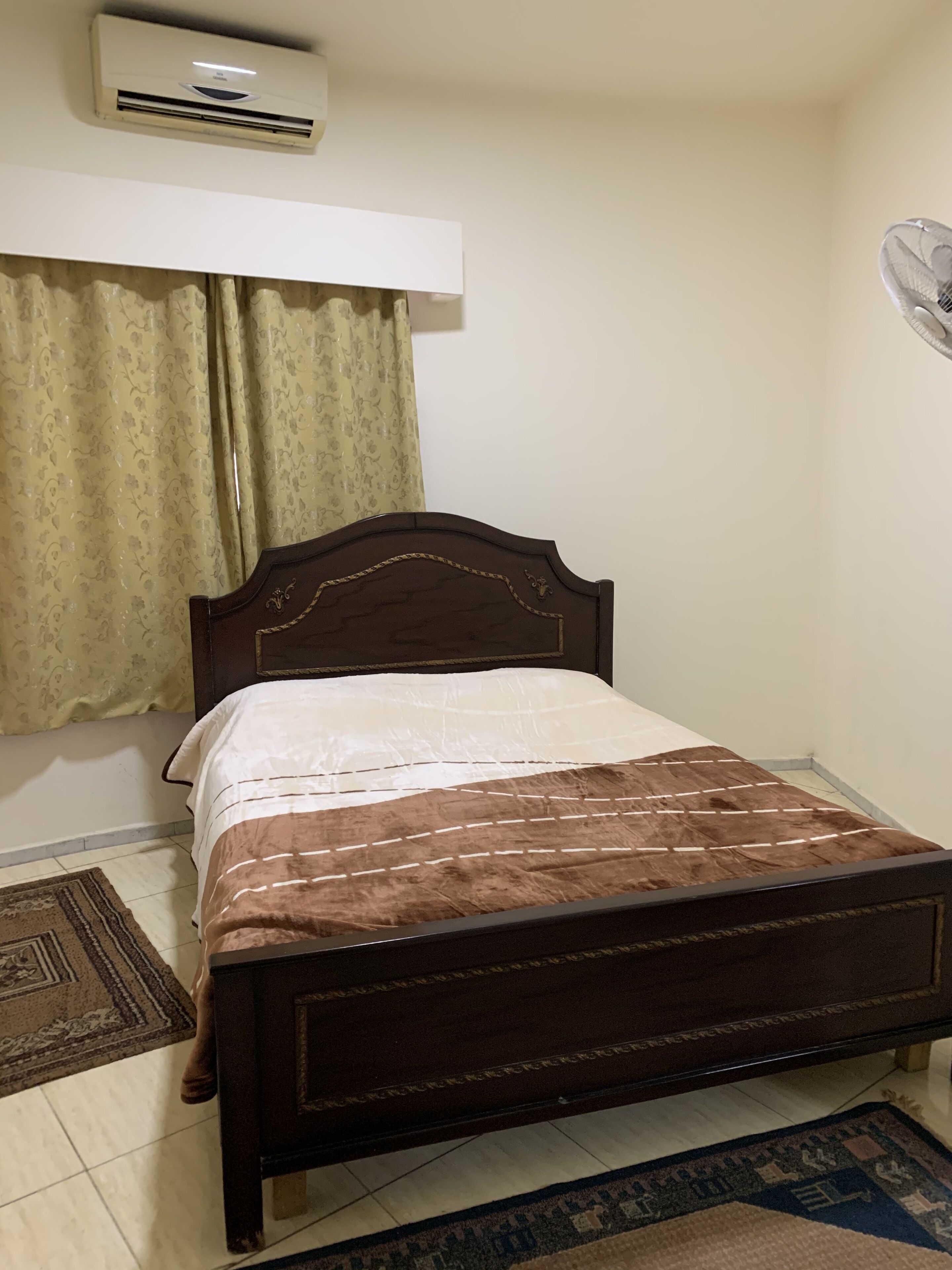 Ein Einzelbett mit Holz-Kopfstück, einer gemusterten Bettdecke und einer braunen Decke. Auf dem Boden liegt ein kleiner Teppich, an der Wand ist eine Split-Klimaanlage angebracht und es gibt einen Vorhang mit Blumenmuster.