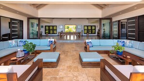 A spacious lounge area with a central dining table, cushioned seating, and large windows offering a view of the outdoors.