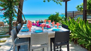 An outdoor dining area with a view of the ocean, featuring a table set with blue and pink tableware, wicker chairs, and a wooden pergola.