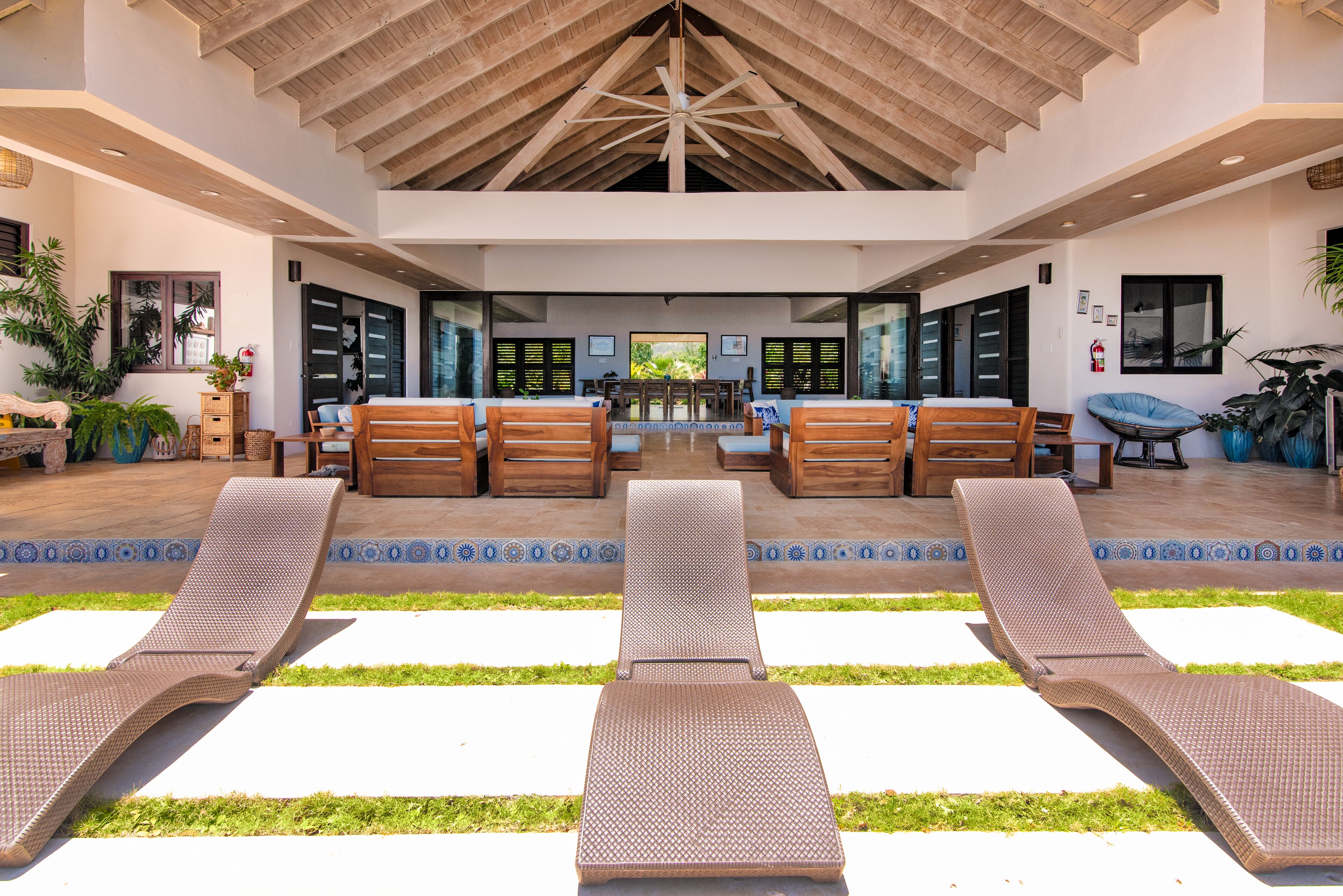 Amplo espaço interno com teto alto e vigas de madeira, com área de estar junto à piscina, incluindo cadeiras de descanso e uma mesa de jantar.