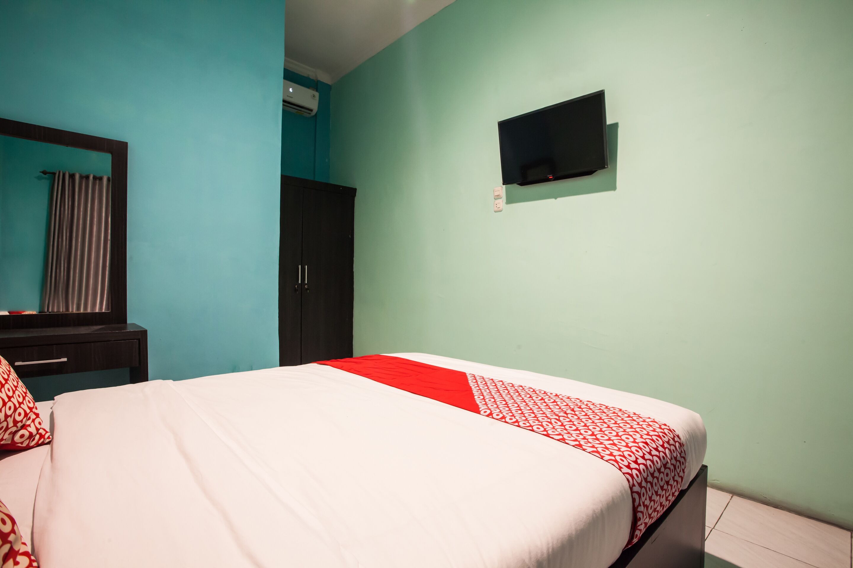 A room with a bed, a dark wardrobe, a flat-screen TV mounted on the wall, and a mirror above the wardrobe.