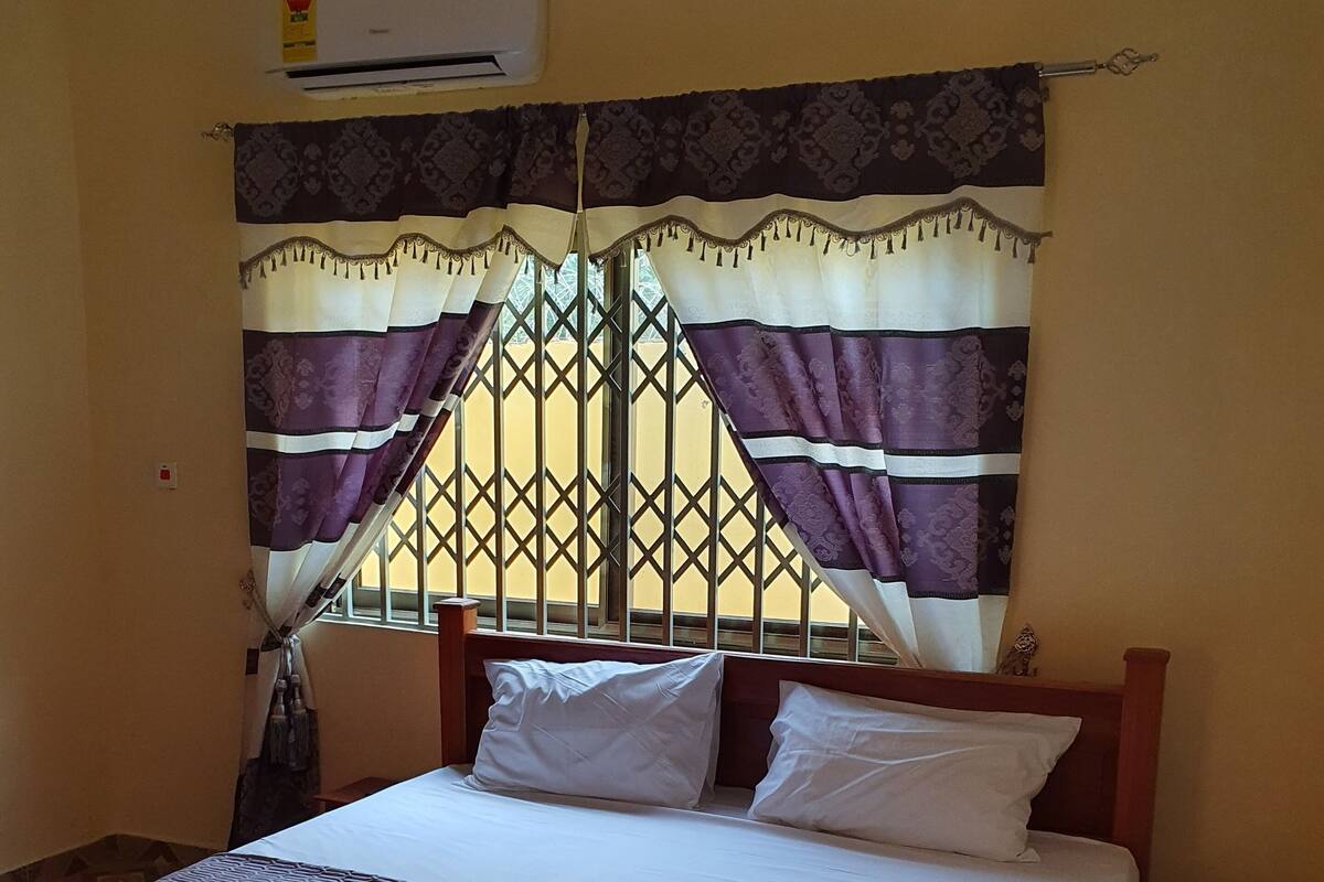 Un lit recouvert d’une couvre-lit à motifs, une fenêtre agrémentée de rideaux décoratifs et un appareil de climatisation placé au-dessus de la fenêtre.