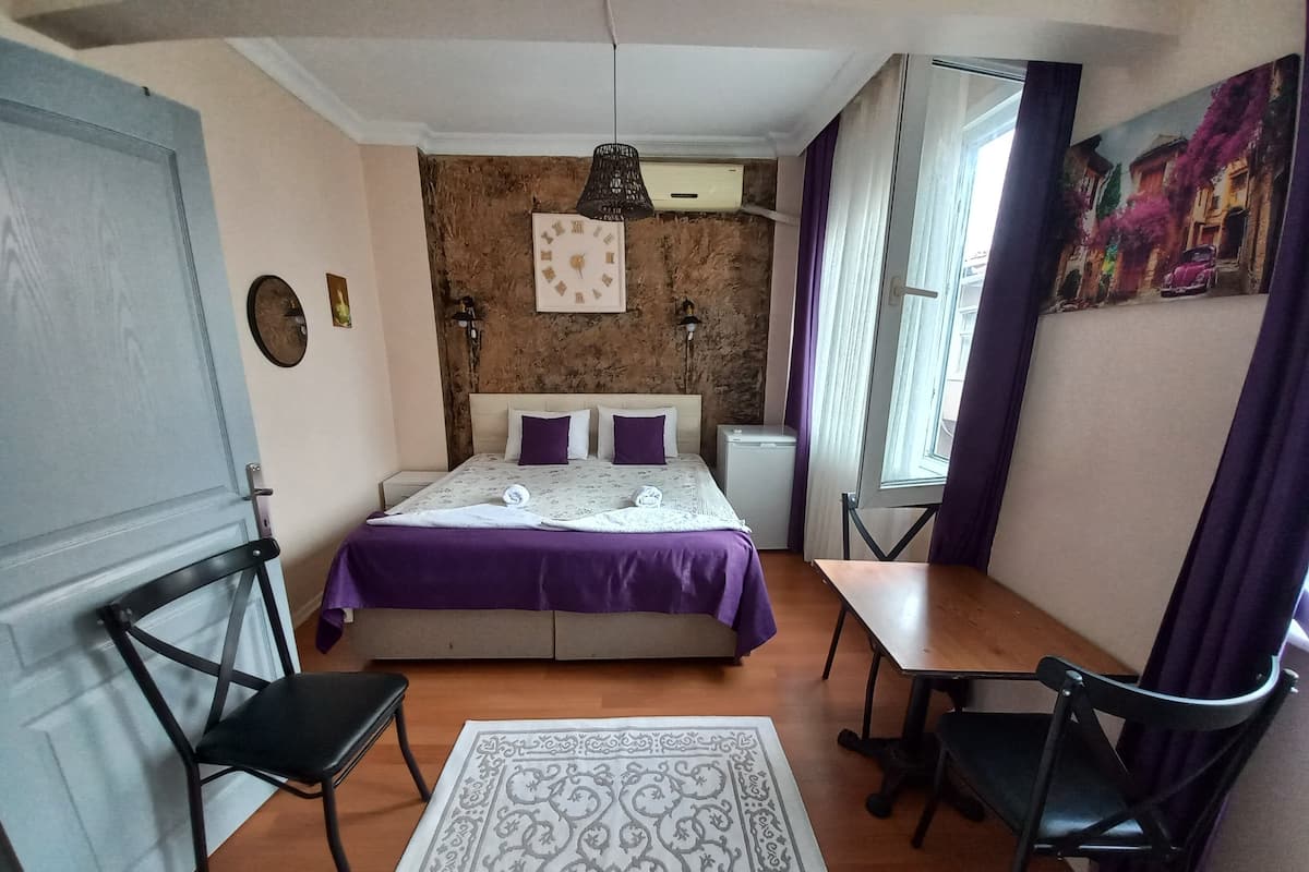 A bedroom with a bed, two chairs, a small table, a wall clock, and a window with curtains.