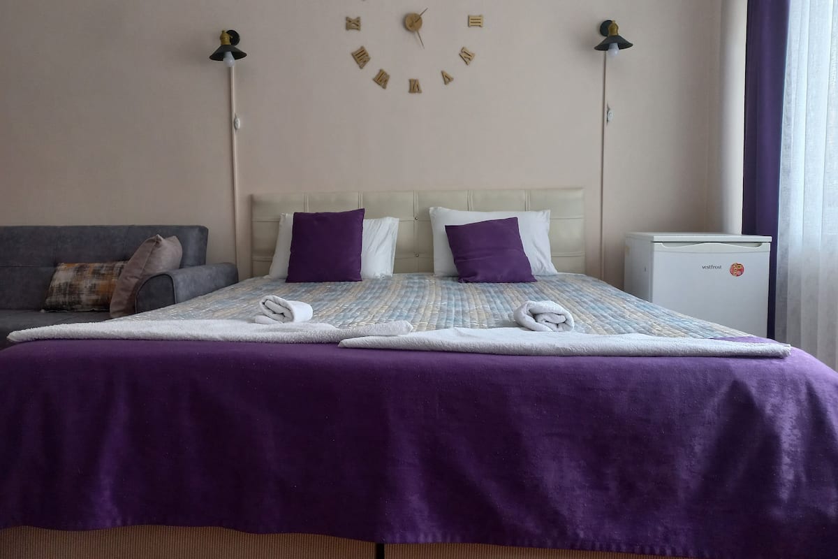 A neatly made bed with a purple blanket, two pillows, and two folded towels. There is a wall clock above the bed, a small white refrigerator to the right, and a grey sofa with cushions to the left.