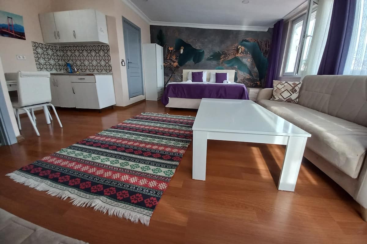 A modern living room with a sofa, a coffee table, a patterned rug, a bed with a purple bedspread, and a kitchen area with cabinets and a refrigerator.