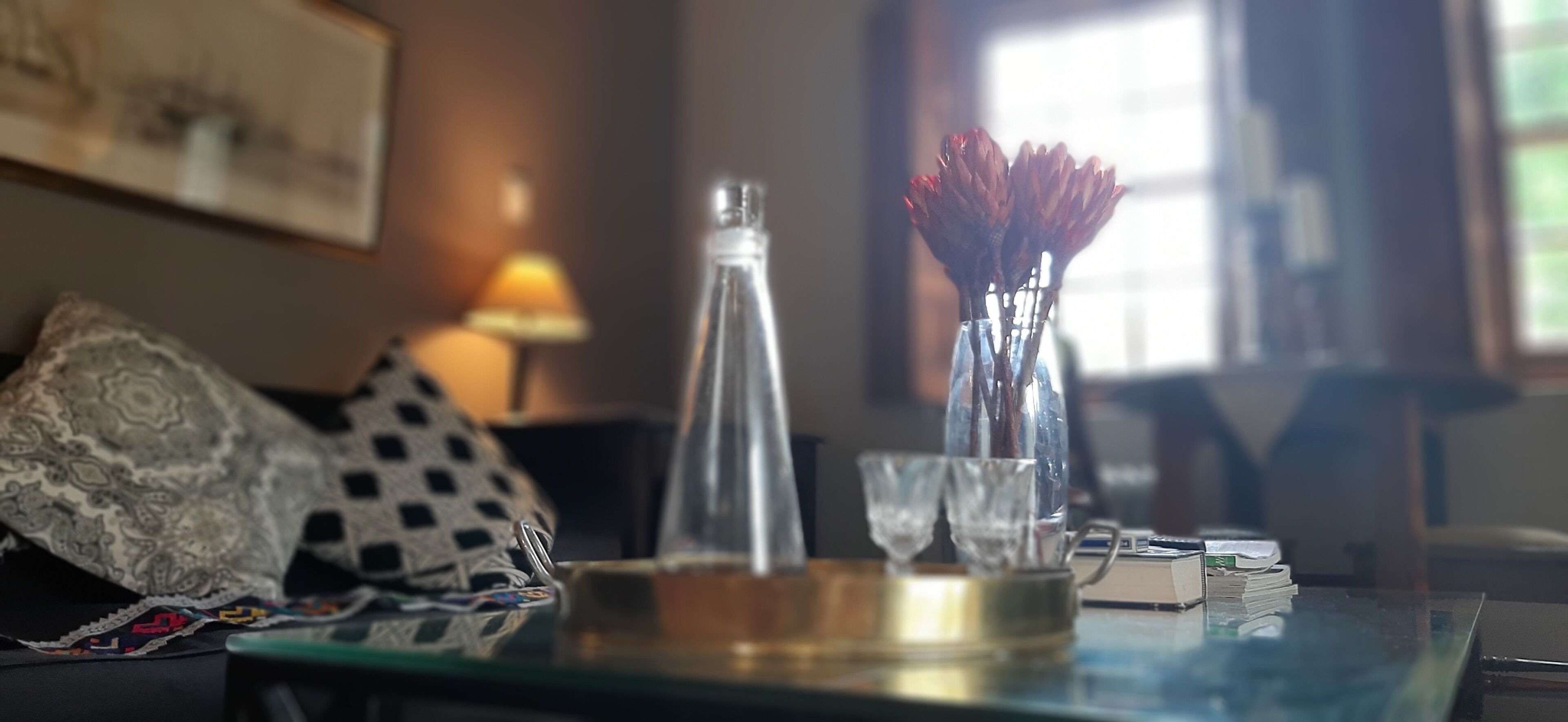 A glass table with a bottle, two vases with dried flowers, and a lamp in a room with a patterned throw pillow and a framed picture on the wall.