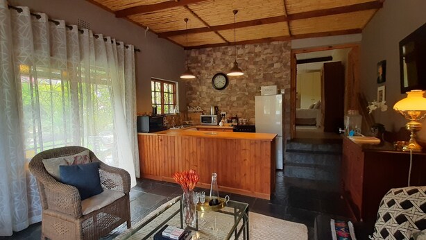 A cozy living room with a wicker sofa, a glass coffee table, and a dining area with a wooden counter. A kitchen is visible in the background with a refrigerator and a clock on the wall. - Abigails Garden Cottage (Montagu)