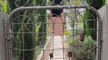 Une entrée fermée par un portail en métal, un chemin menant à un bâtiment et une végétation luxuriante tout autour.