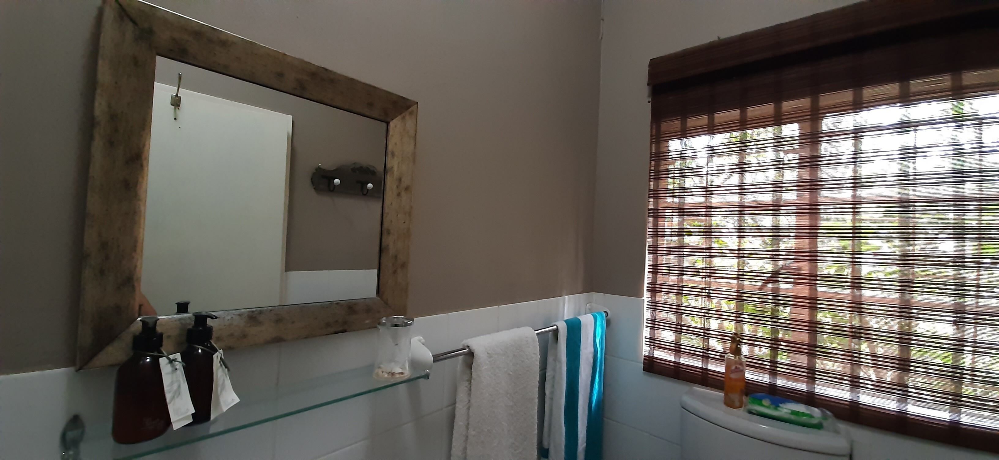 Ein Badezimmer mit einem spiegelnden Holzbettrahmen, einer Glasablage mit Pflegeprodukten, einem Handtuchhalter mit einem gestreiften Handtuch und einem Fenster mit Jalousien.