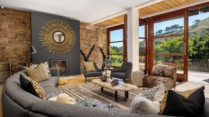 A modern living room with a stone wall, a fireplace, and a large window with a view of greenery. - Le Thallo (Cape Town)