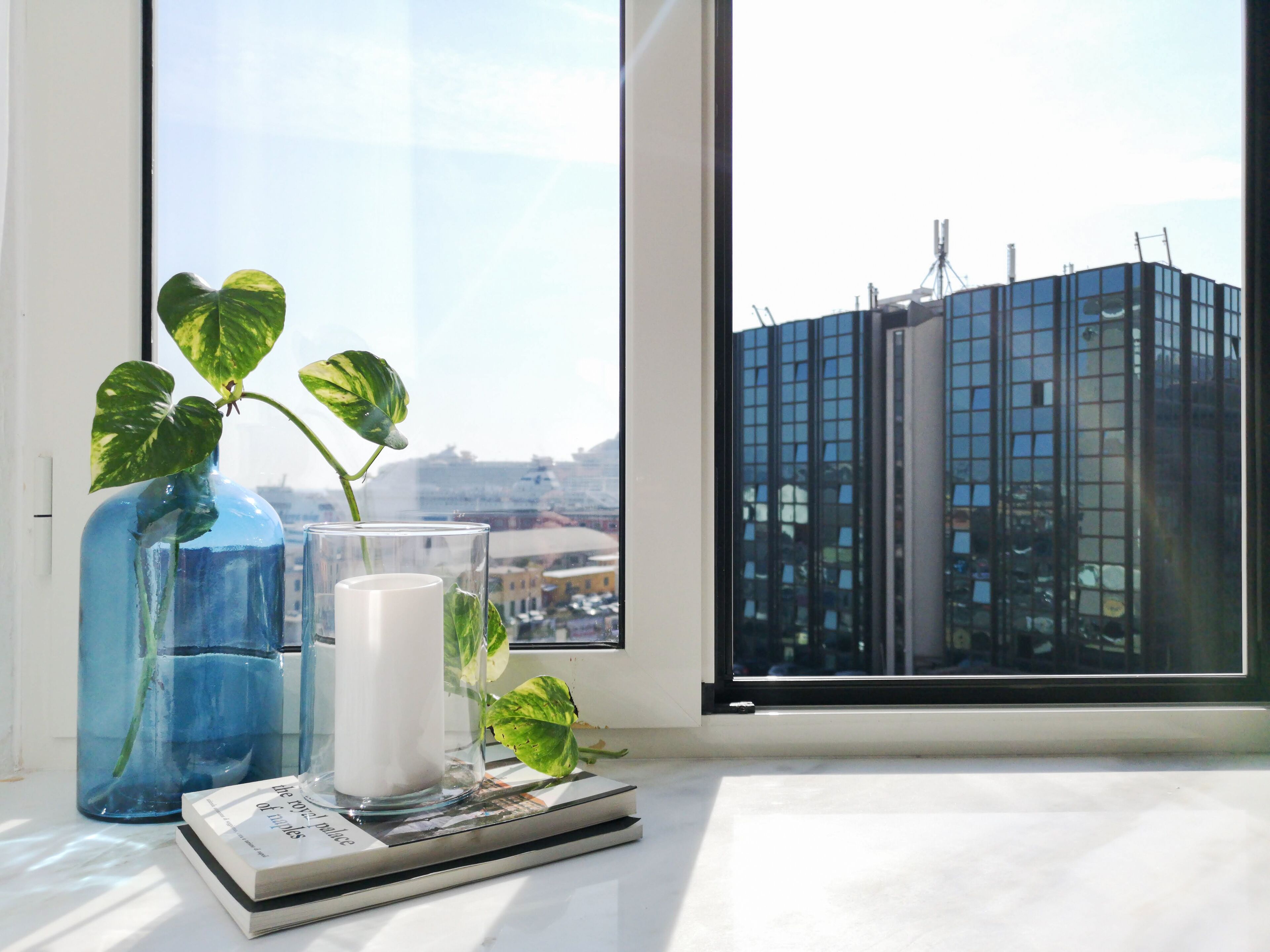 A window with a view of a modern building, a blue vase with a plant, and a candle on a windowsill.