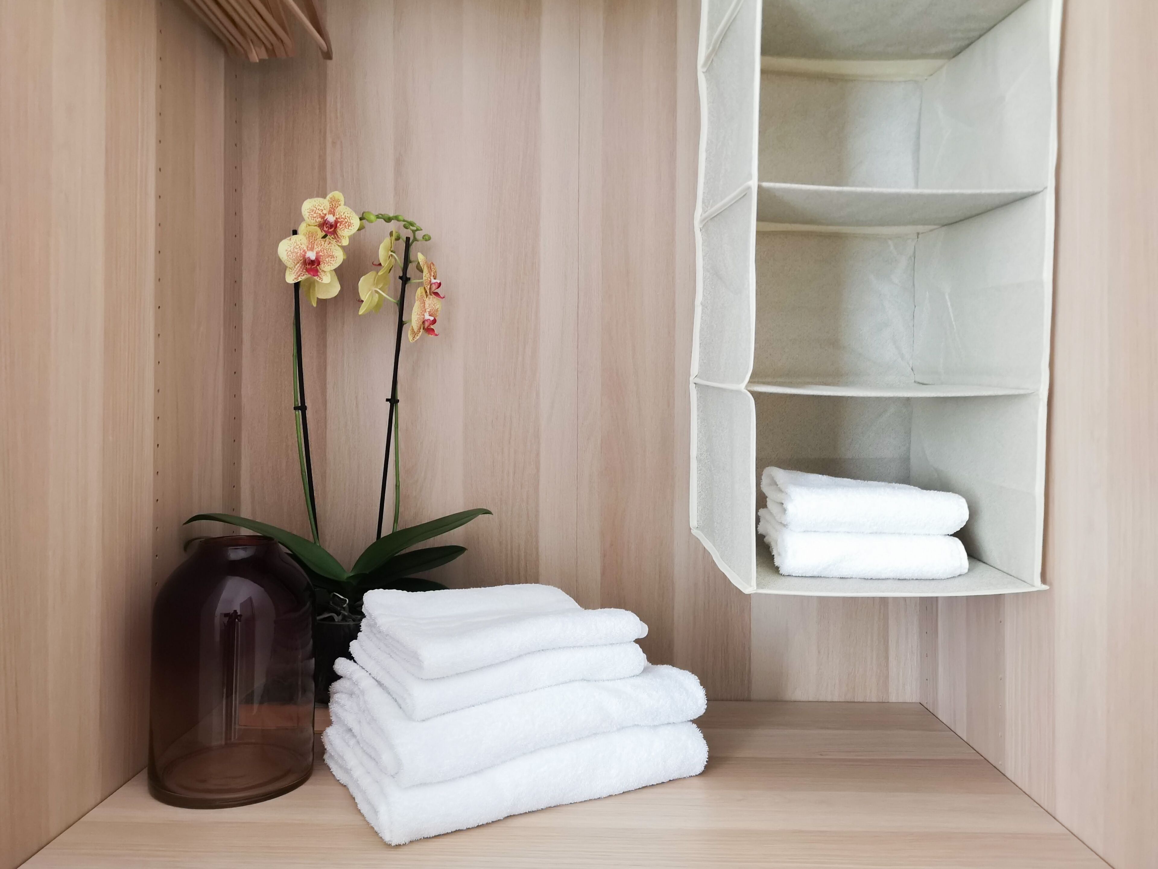A corner of a room with a wooden wall, a glass vase with a plant, and a white shelf with folded towels.