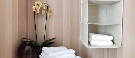A corner of a room with a wooden wall, a glass vase with a plant, and a white shelf with folded towels.