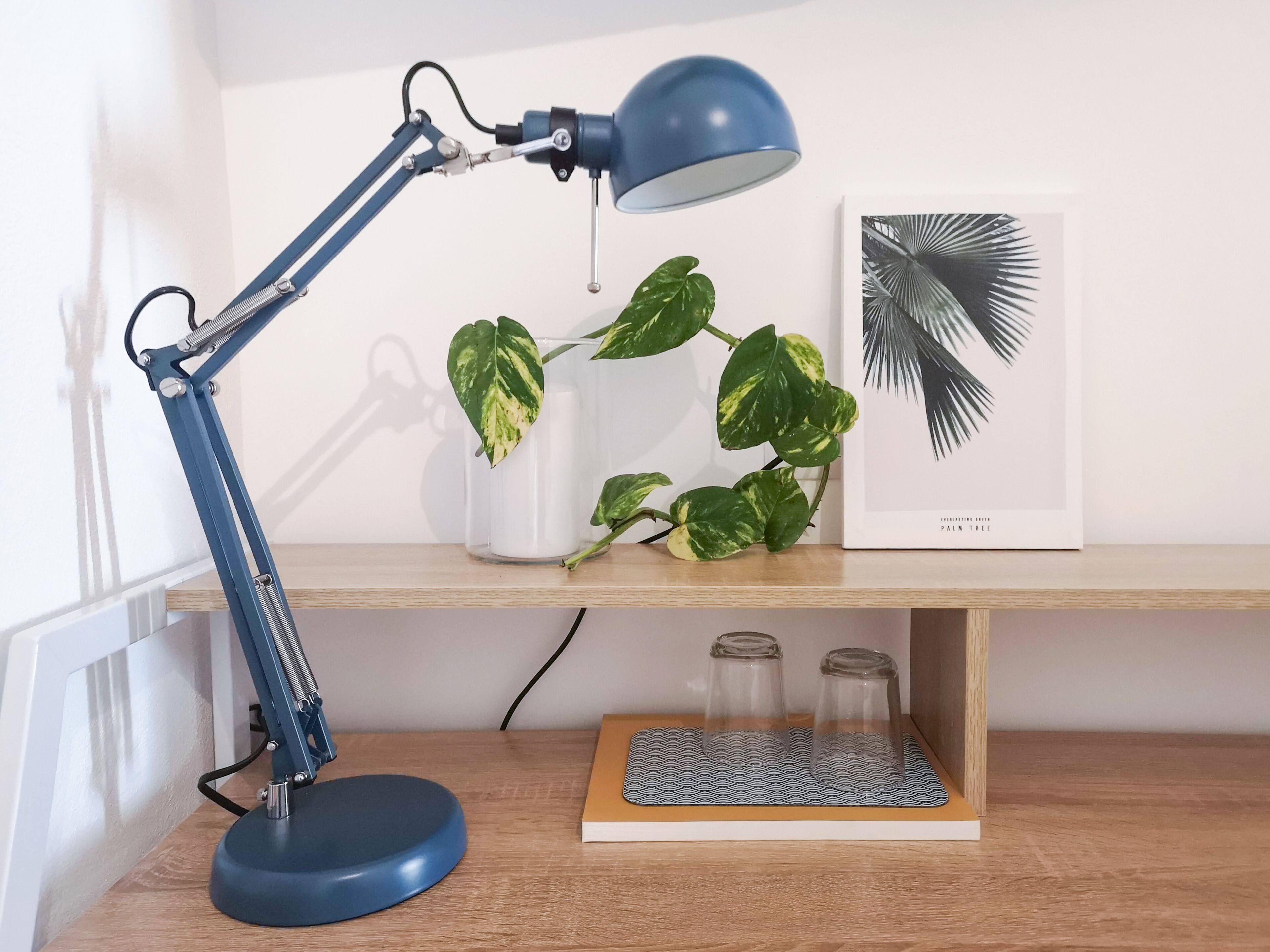 A desk with a blue desk lamp, a potted plant, a framed picture, and a keyboard on a wooden surface.