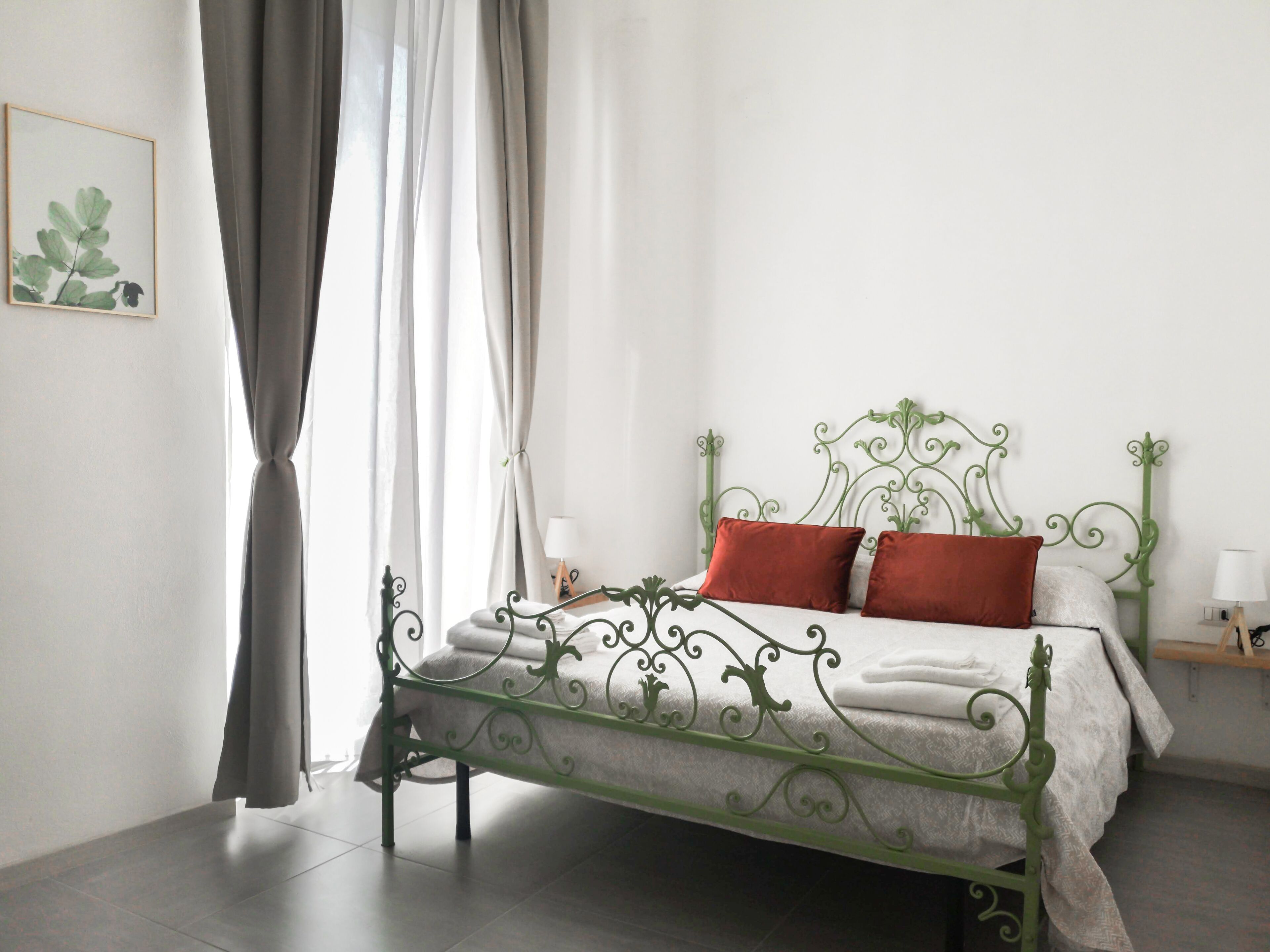 Un dormitorio con estructura de cama de metal verde, ropa de cama blanca y cortina gris.