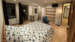Un dormitorio con cama, una silla, un televisor, un espejo y un baño con ducha y lavamanos.