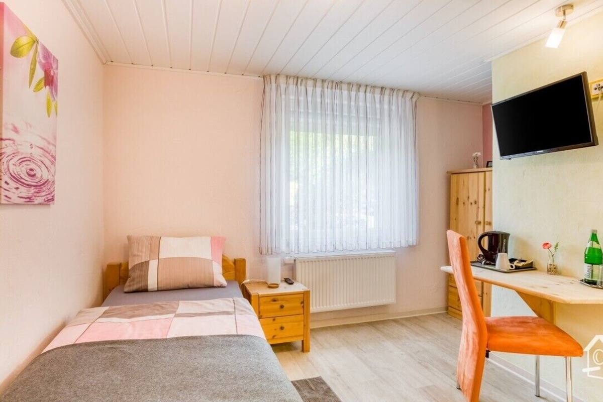 Ein Schlafzimmer mit Bett, Schreibtisch, Stuhl, Fernseher und einem Fenster mit Vorhängen.