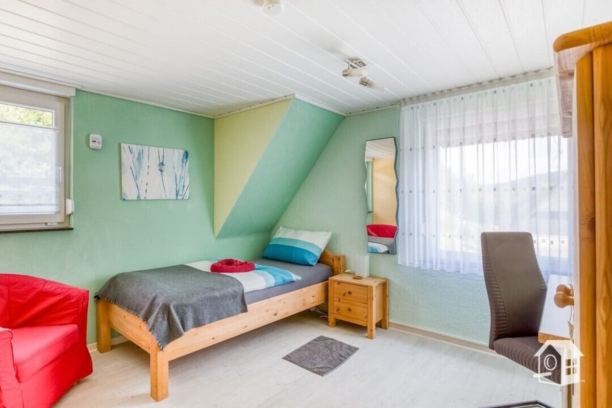 Ein Zimmer mit einem Bett, einem roten Sessel, einem Nachttisch aus Holz, einem Spiegel und einem Fenster mit Vorhängen.