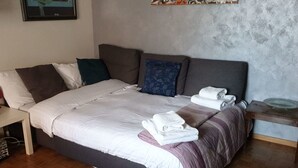 Una cama bien hecha con sábanas blancas y una manta morada, una mesita de noche de madera con un objeto de vidrio, y un cabecero gris.