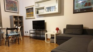 Una sala de estar con piso de madera, un sofá gris, un televisor, una mesita blanca y una lámpara roja.