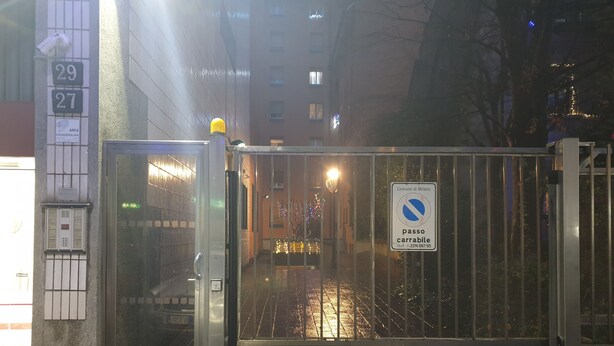 Una entrada protegida por un portón metálico, un cartel que indica zona sin estacionamiento y un edificio con ventanas numeradas al fondo.