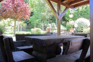 An outdoor patio with a wooden table and benches, surrounded by lush greenery and trees.