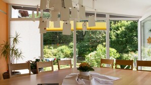 Comedor con mesa de madera, sillas, planta en maceta y vistas a un jardín exuberante a través de amplios ventanales.