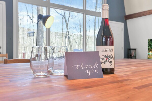 A bottle of Cherry Blossom Pinot Noir, two empty glasses, and a thank you card on a wooden table with a window in the background. - The Reed – A Cozy Cabin in the Woods (Rockbridge)