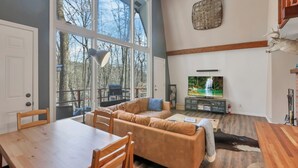 A spacious living room with a large window, a wooden dining table, a sofa, and a television. - The Reed – A Cozy Cabin in the Woods (Rockbridge)