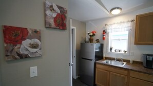 Cocina compacta con refrigerador, pileta y alacenas de madera. Hay cuadros con motivos florales en las paredes y una ventana con cortina.