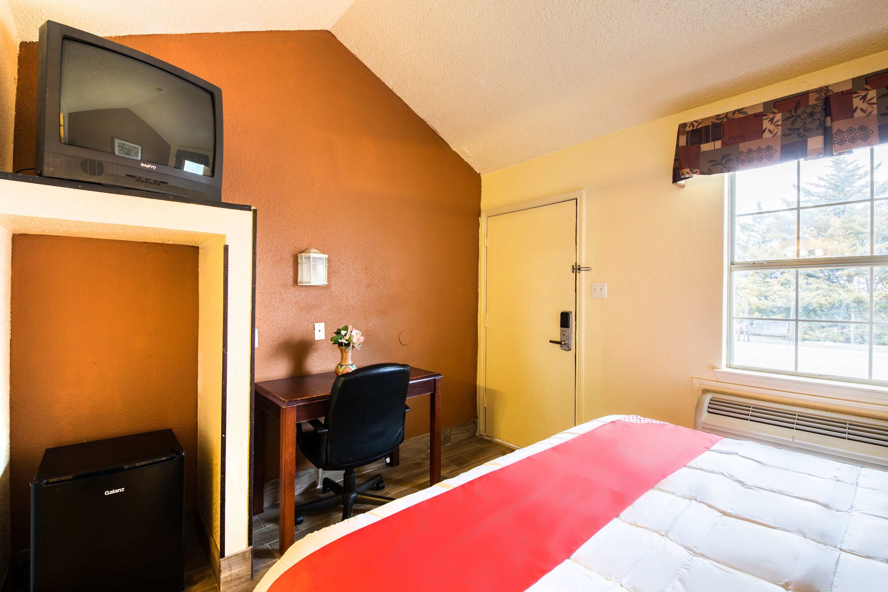 Habitación de hotel con cama, escritorio, silla, televisor y puerta.