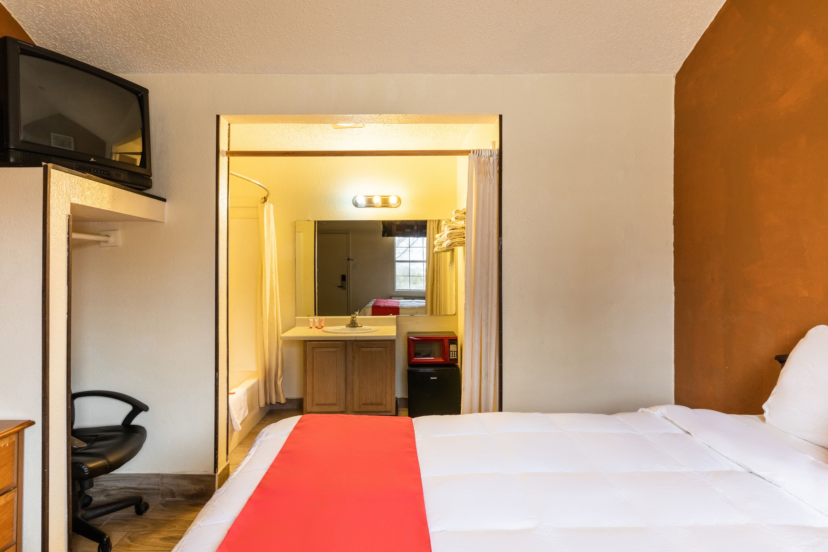 A hotel room with a bed, a desk with a chair, a TV, a bathroom with a sink and mirror, and a red blanket on the bed.