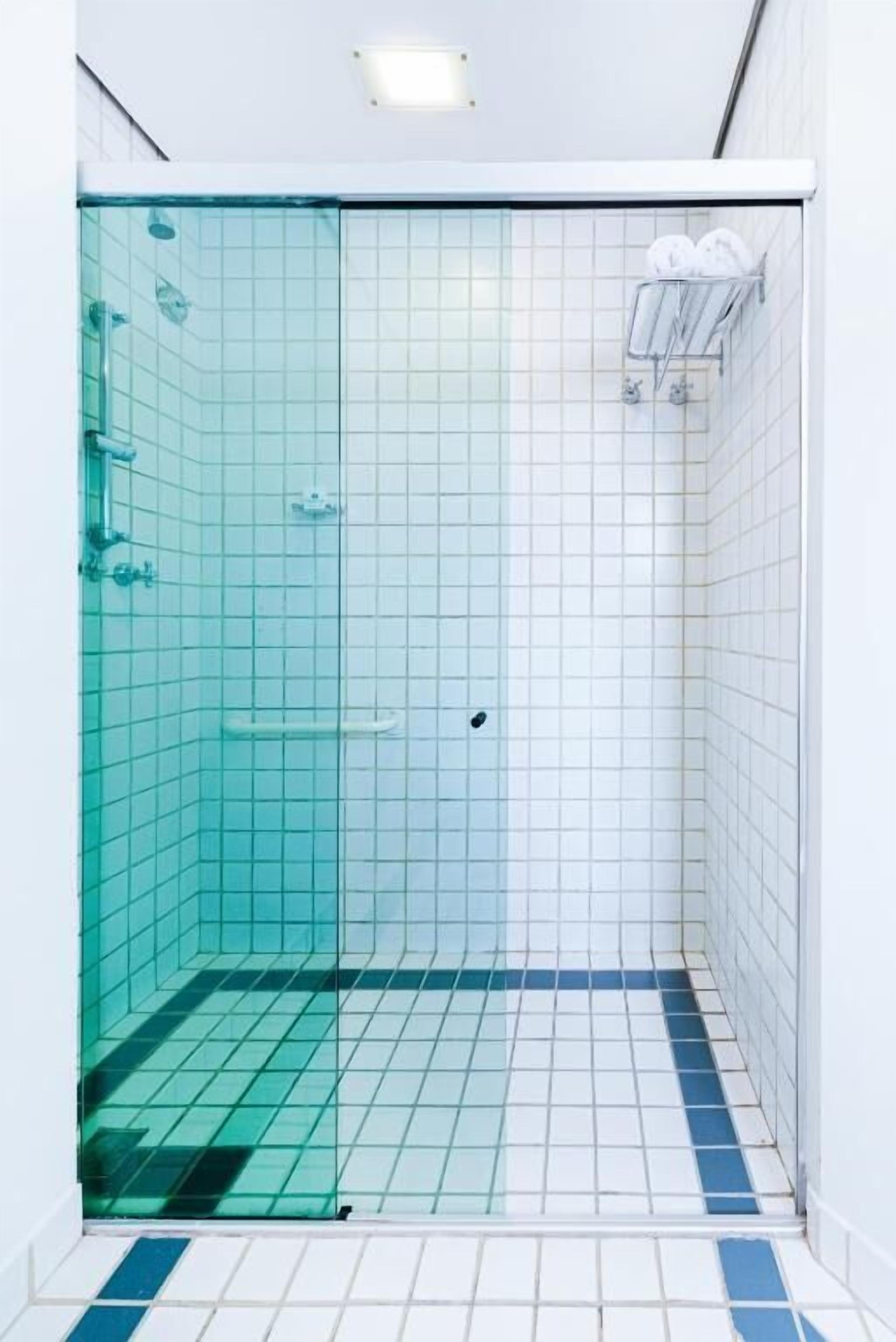 Une douche avec une porte en verre, des murs recouverts de carreaux blancs et une bande bleue sur le sol.