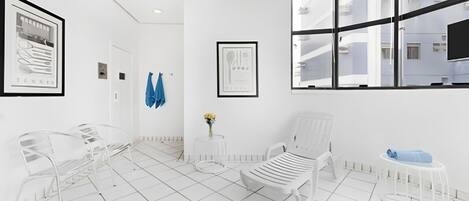 A modern, white interior with a large window, a white chair, a small table with a vase of flowers, and a framed picture on the wall.