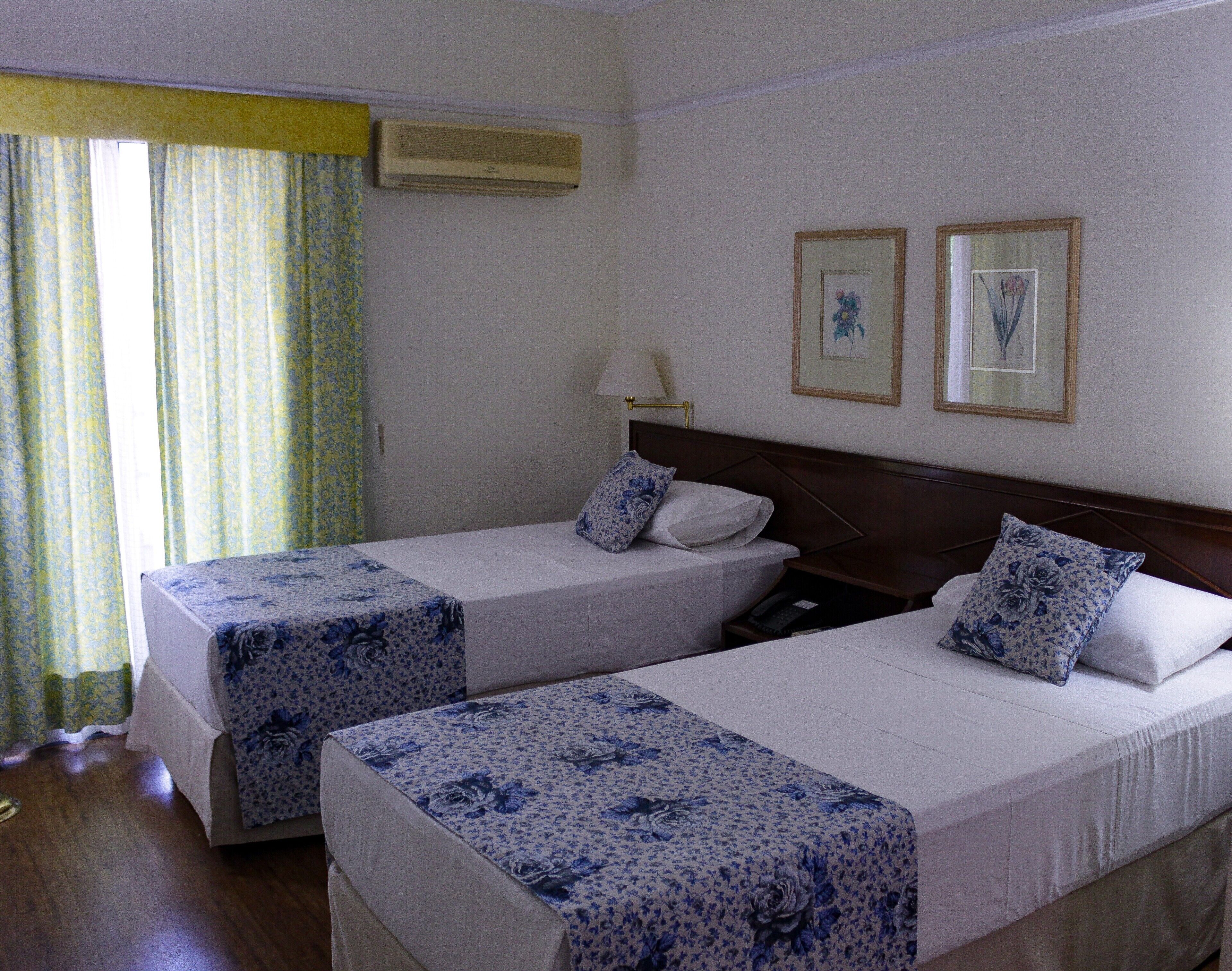 A hotel room with two beds, a nightstand, a lamp, and a window with curtains.
