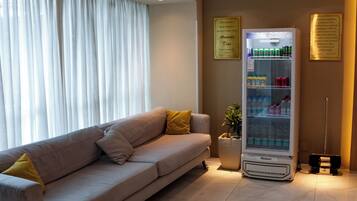 Quarto de hotel moderno com sofá bege, minibar e uma grande janela com cortinas brancas.