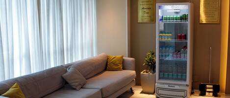 A modern hotel room with a beige sofa, a minibar, and a large window with white curtains.