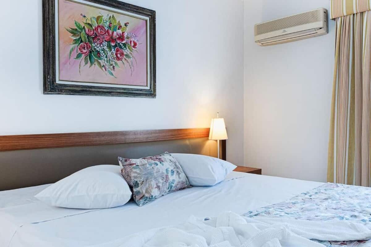 Une chambre d’hôtel comprenant un lit, une œuvre d’art florale encadrée, une lampe de chevet, un appareil de climatisation et une fenêtre avec des rideaux.