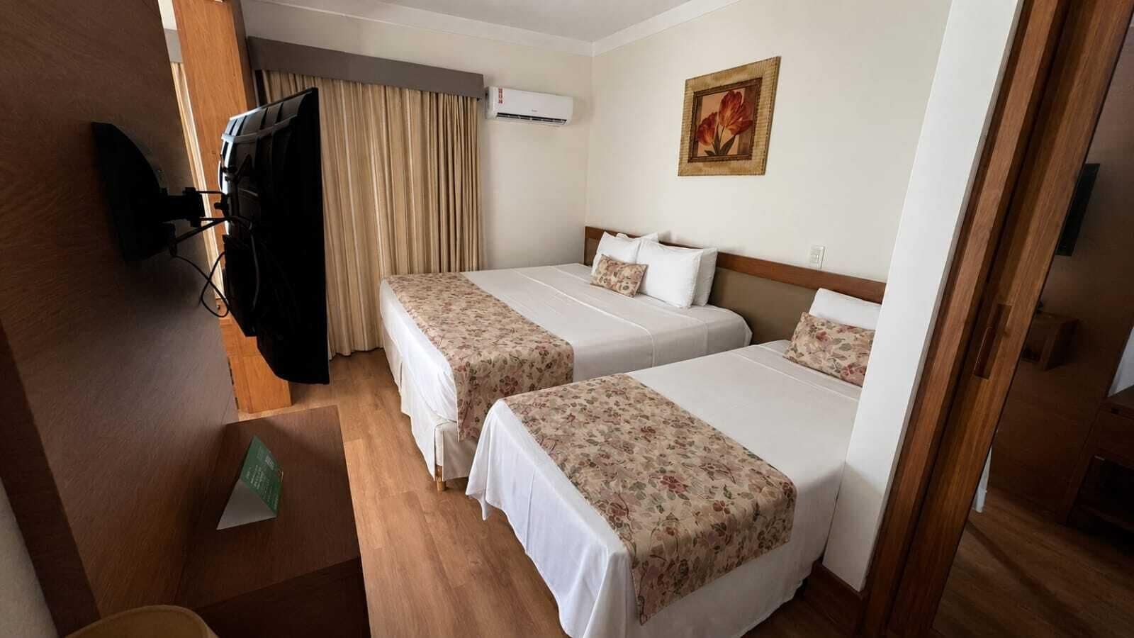 A hotel room with two beds, a television, an air conditioning unit, and a painting on the wall.