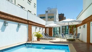 Una piscina con sillas de descanso y sombrillas, rodeada por un muro de ladrillo y puertas de cristal.