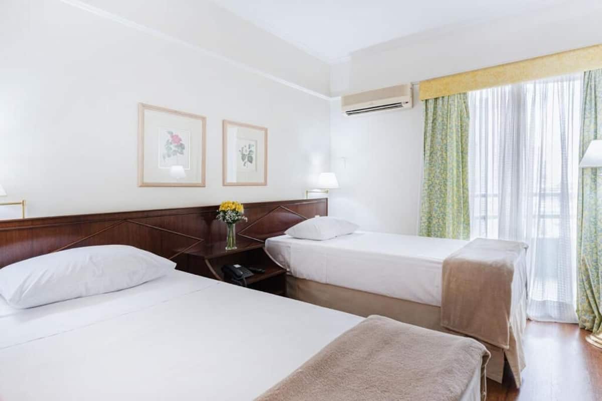 Une chambre d’hôtel avec deux lits simples, une table de chevet avec une lampe, des tableaux encadrés au mur et une fenêtre avec des rideaux.