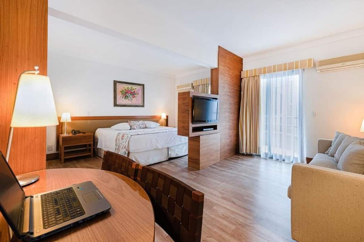 Une chambre d’hôtel avec un lit, un bureau avec un ordinateur portable, une télévision, un canapé et une fenêtre avec des rideaux.