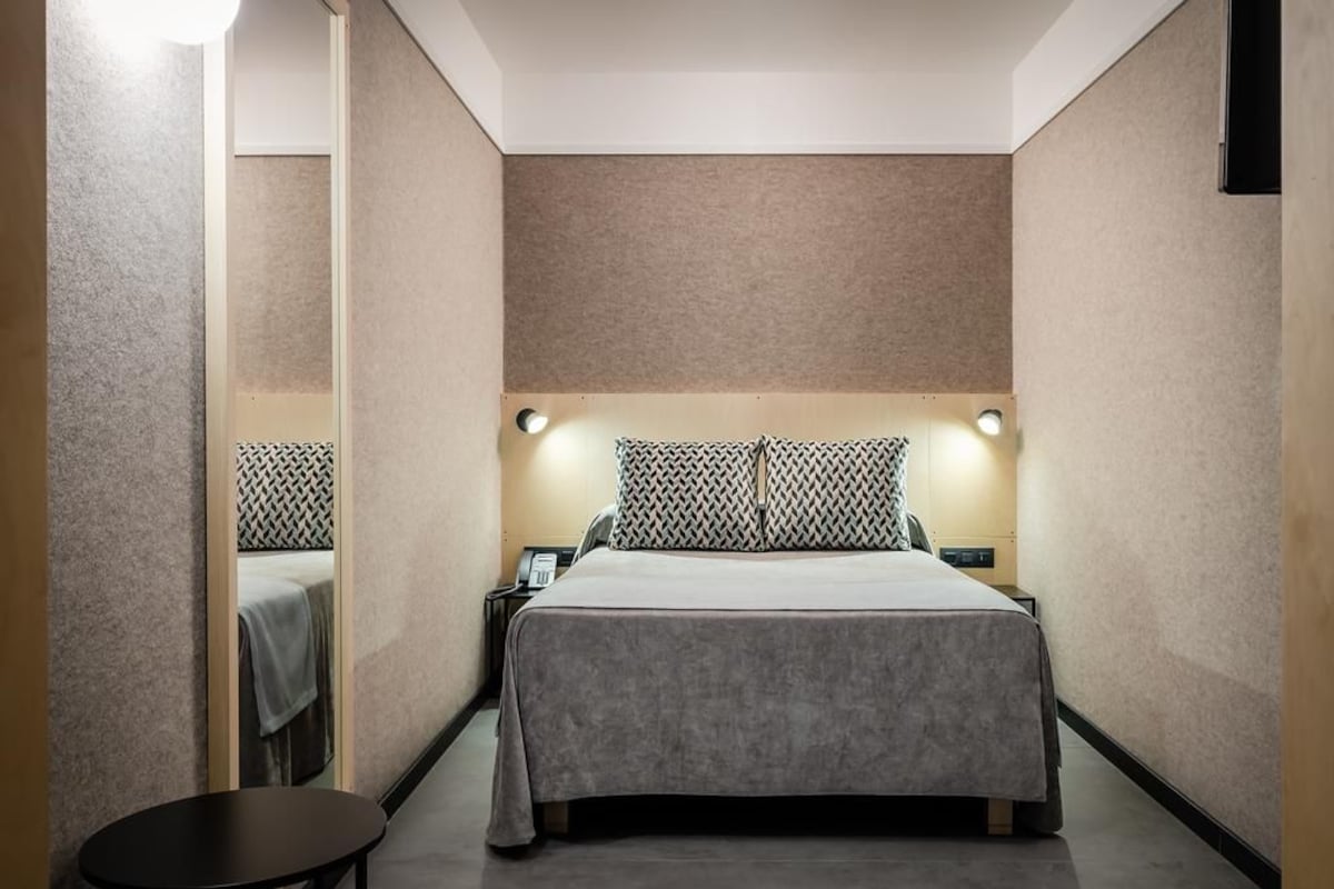 Una habitación de hotel con cama, mesitas de noche, una pequeña mesa y una lámpara fijada en la pared.