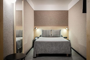Una habitación de hotel con cama, mesitas de noche, una pequeña mesa y una lámpara fijada en la pared.