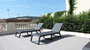 Terraza en la azotea con dos tumbonas y una mesa, rodeada de vegetación y un edificio blanco con chimenea.
